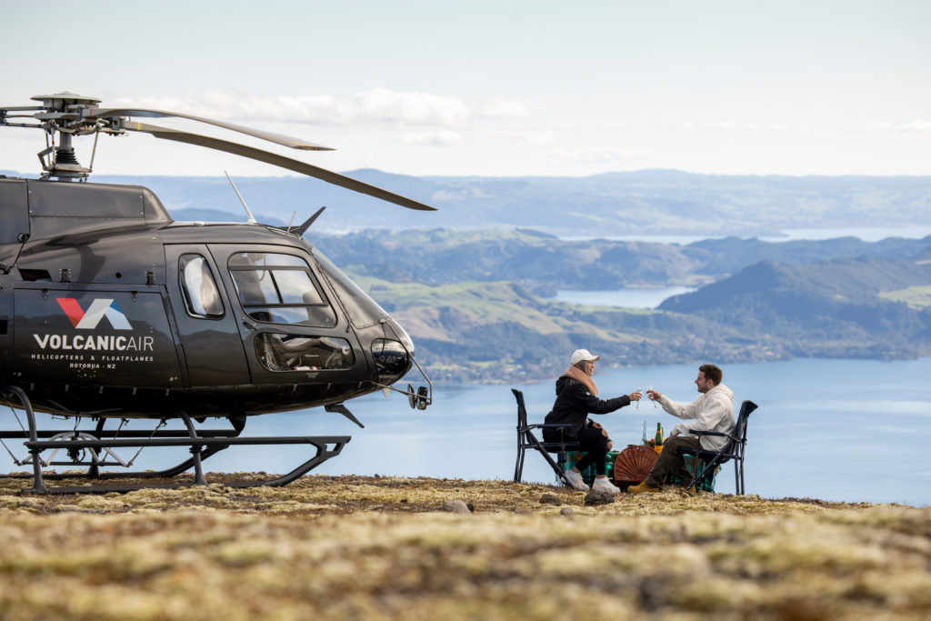 Man and woman cheersing on top of mountain next to a helicopter